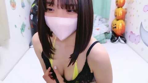 Media: Video of an Asian woman with straight black hair, wearing a yellow bra and a pink face mask, standing in a Halloween-themed room with orange pumpkins.