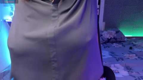 Media: A video of a person wearing a light grey, button-up shirt, partially obscured by a blue light, standing in a dimly lit room with a bed and floral-patterned blanket in the background.