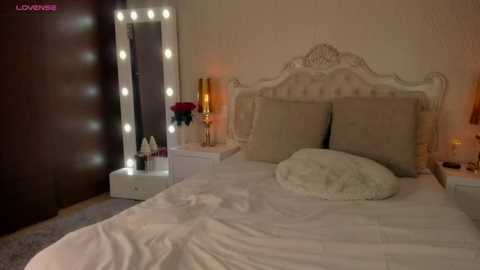 Media: Video of a cozy, dimly lit bedroom with a white, ornate headboard, white bedding, and a fluffy white pillow. A vanity mirror with a string of lights reflects the room, and a nightstand holds a gold lamp, a bouquet of red roses, and makeup items.