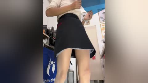 Media: Video of a woman in a black skirt, white blouse, and beige tights, taken from a low angle. Background shows a cluttered office with a blue banner and whiteboard.