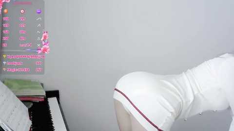 Media: A video showing a woman in a white, tight, sleeveless dress bending over a black piano in a bright, minimalist room with a pink flower arrangement.