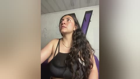 Media: Video of a Latina woman with long, wavy black hair, wearing a black crop top, standing in a dimly lit room with a purple guitar leaning against a white wall.