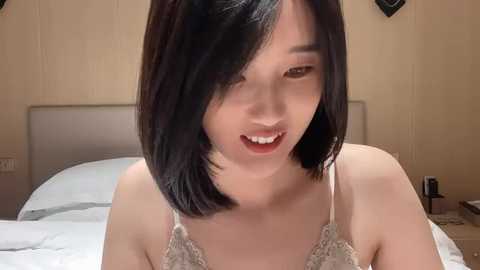 Media: Video of an East Asian woman with shoulder-length black hair, fair skin, and a slender physique. She wears a lacy, off-white bra. Background features a beige wall, a bed with white sheets, and a nightstand with a lamp.