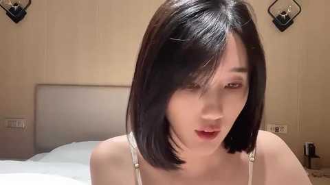 Media: Video of an Asian woman with straight, shoulder-length black hair and fair skin, wearing a white spaghetti-strap top, in a beige-walled bedroom with a bed and two wall sconces.