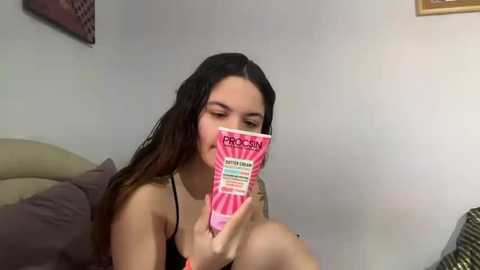 Media: Video of a young woman with long, dark hair, reading a pink book titled \"Pregnancy.\" She wears a black tank top, sitting on a beige couch in a dimly lit room with gray walls.