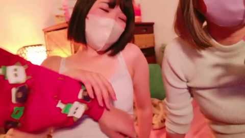 Media: A video of a young woman with a white mask and bob haircut, wearing a white tank top, hugging a plush toy. A second woman in a beige sweater is also masked, standing beside her. Background features a wooden dresser and a cozy, warm room setting.