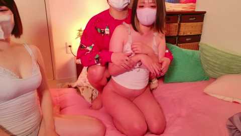 Media: Video of three Asian women in a bedroom, two kneeling on a pink bed, wearing masks, one topless, the other in a white bra, surrounded by green and white pillows.