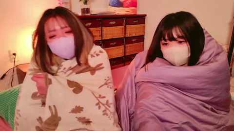 Media: Video of two Asian women wearing face masks, wrapped in blankets, sitting on a bed in a cozy, warmly lit room with a dresser and potted plants in the background.