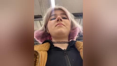 Media: Video of a young woman with short, dyed pink hair, wearing a black choker and a mustard-yellow jacket over a black top. The background shows a white ceiling and pipes.