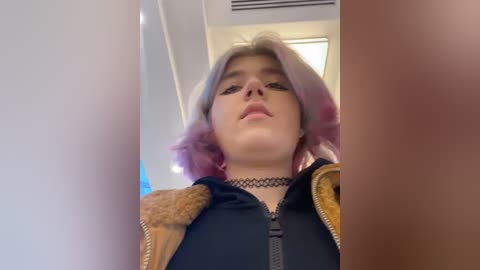 Media: Video of a young person with pink hair, wearing a black choker, a brown jacket, and a black hoodie, taken from a low angle, indoors.