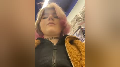 Media: Video of a young woman with pastel pink hair and a black choker, wearing a black zip-up jacket, captured from a low angle in a cluttered room with blue and white decor.