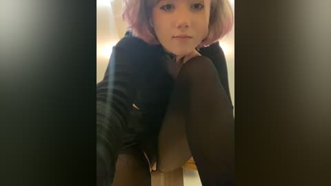 Media: A video of a young woman with short, dyed pink hair, wearing a black leather jacket and black tights, sitting on a wooden floor, looking directly at the camera.