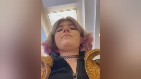 Media: Video of a young woman with pink hair, wearing a black choker, a brown jacket, and looking down. The background shows a white ceiling and window.