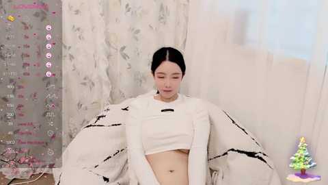 Media: Video of a fair-skinned East Asian woman with dark hair, wearing a white crop top and a large, decorative white cape with black trim, sitting in a room with light floral-patterned curtains and a small Christmas tree on the right.