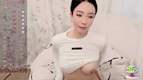 Media: A video of an East Asian woman with fair skin, black hair, and small breasts, lifting her white shirt to expose her chest, sitting indoors with floral curtains and a Christmas tree in the background.