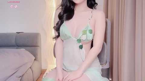 Media: Video of a fair-skinned, long-haired Asian woman in a delicate, mint-green lingerie dress with floral patterns, seated on a gray couch in a modern bedroom.