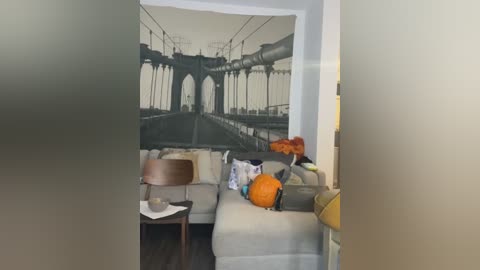 Media: A video of a cozy living room featuring a large, wall-mounted mural of the Brooklyn Bridge. The room includes a beige sofa, a wooden chair, and a modern lamp on a small wooden table.