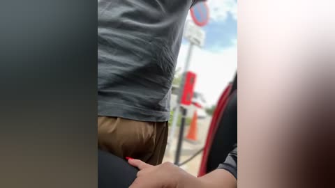 Media: Video of a man's torso in a grey T-shirt and brown pants, leaning over a car seat, with a woman's hand holding his arm, in a car with a red interior.