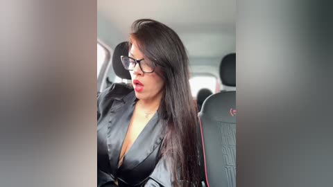 Media: A video of a Latina woman with long, straight dark hair, wearing black glasses and a black satin blouse, sitting in a car. The background features a blurry car interior.