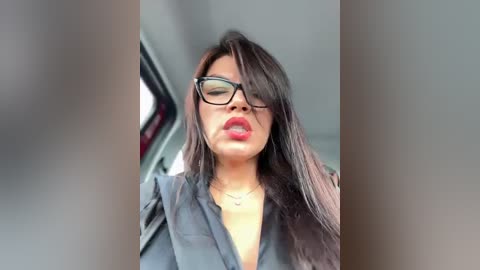 Media: Video of a woman with long, straight black hair, wearing black-framed glasses, red lipstick, and a black shirt, captured from a car's rearview mirror.