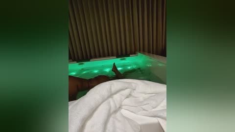 Media: Video of a person in a jacuzzi with green underwater lighting, wearing white towels, and lying against a beige wall with vertical slats.