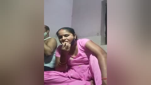 Media: A video shows a woman in a pink sari sitting on a bed, covering her mouth, and a man in the background. The room has light purple walls and a white bedspread.