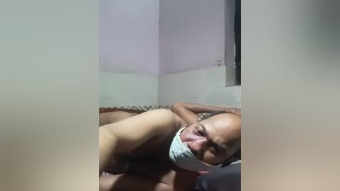 Media: Video of a bald, muscular, nude man with a light skin tone, wearing a surgical mask, lying on a bed with a patterned quilt. The room has tiled walls and a dim, indoor lighting setup.