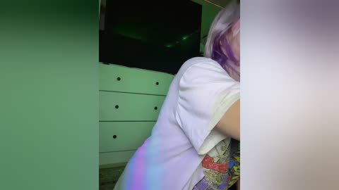 Media: A video of a person with long, pastel-colored hair, wearing a white t-shirt with a colorful pattern, standing in a dimly lit room with a green tint. The background features a white dresser and a partially visible TV.