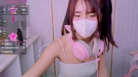 Media: A video of an Asian woman with long dark hair, wearing a pink face mask, white tank top, and pink headphones, sitting in a modern room with a white counter and a star filter overlay.