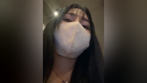 Media: Video of a woman with long black hair, wearing a white face mask and a black top, indoors with blurred background.