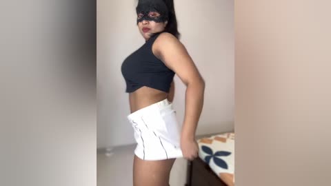 Media: Video of a slender woman with medium skin tone and dark hair, dressed in a black mask, black crop top, and white high-waisted shorts. She stands indoors against beige walls and a bed with a floral-patterned blanket.