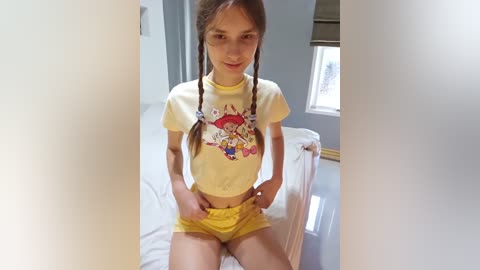 Media: Video of a young girl with light skin, brown braids, and a yellow T-shirt with cartoon characters, wearing yellow shorts, sitting on a bed in a minimalist room with white walls and a window.