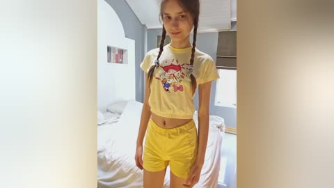 Media: Video of a young woman with light skin, long brown hair in pigtails, wearing a yellow crop top and shorts, standing in a bright, modern bedroom with a bed, white walls, and a window.