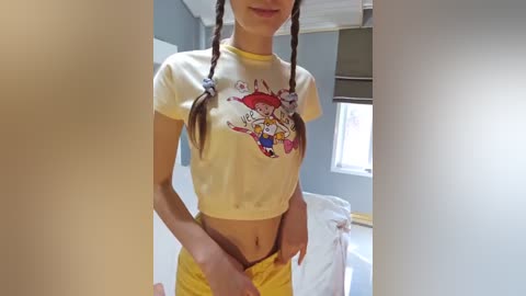 Media: Video of a slender, fair-skinned woman with long braided hair wearing a yellow crop top and yellow shorts, standing in a simple, gray-painted room with a window and bed visible.