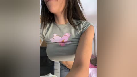 Media: Video of a young woman with light skin and long brown hair, wearing a tight, light gray crop top with a pink heart graphic and low-rise jeans. Background shows a dimly lit bedroom with a bed and a dark dresser.