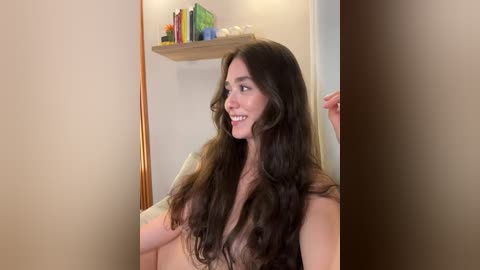 Media: Video of a young woman with long, wavy dark hair, fair skin, and a slender physique, smiling, standing in a dimly lit room with beige walls and a wooden shelf holding colorful books and a green plant.