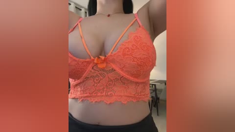 Media: Video of a light-skinned woman wearing a lace orange bra with thin straps, revealing medium-sized breasts. Background shows a blurred, modern living room with a white lampshade and furniture.