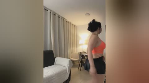 Media: A video captures a woman in a bright orange sports bra and black shorts, standing in a dimly lit, modern hotel room with beige curtains, a white bed, and a desk with a lamp.