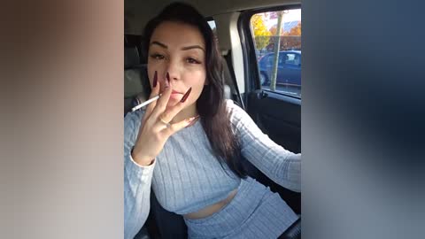 Media: Video of a young woman with long dark hair, wearing a light blue ribbed crop top and matching high-waisted skirt, smoking a cigarette in a car, sunlight filtering through the window.