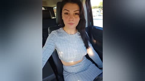 Media: Video of a young woman with long dark hair, wearing a light gray ribbed crop top and matching high-waisted skirt, seated in a car with sunlight streaming in.