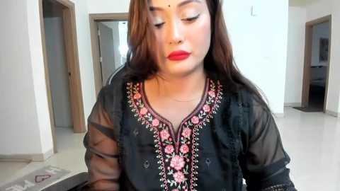 Media: Video of a South Asian woman with fair skin, long dark hair, and red lipstick, wearing a black embroidered shalwar kameez, indoors with white walls and open doors.