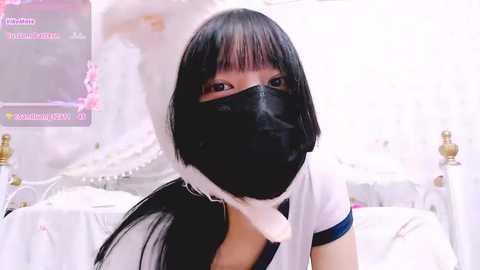 Media: A video of an Asian woman with a black mask, short black hair, wearing a white outfit with blue trim, sitting on a white bed. The background features white bedding and a soft, pastel-colored room.