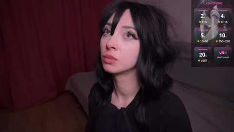 Media: A video of a young woman with long, wavy black hair, light skin, and a slender physique, wearing a black top, looking contemplative. The background features a dark red curtain and a digital screen displaying live stream stats.