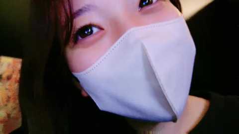 Media: A close-up video of a young Asian woman with fair skin, wearing a white face mask, showing her large, expressive brown eyes. The background is dark, possibly indoors.