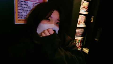 Media: A video shows a person with long dark hair and a white mask partially covering their face, standing in a dimly lit room with a vending machine displaying colorful manga covers.