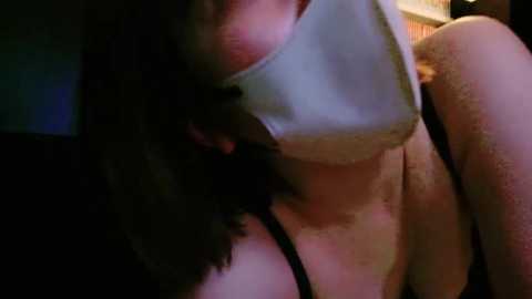 Media: A video of a woman with shoulder-length dark hair, wearing a white mask and a black bra, partially covered by her hair, in dim lighting.