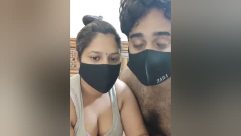 Media: Video of a woman with dark hair in a bun, wearing a black face mask and gray tank top, standing beside a man with curly hair and a black face mask, in a dimly lit room.