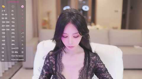 Media: Video of an East Asian woman with long black hair, wearing a black lace robe, sitting on a white couch in a modern living room.