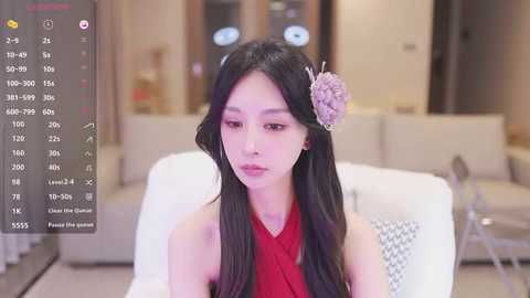 Media: Video of a young East Asian woman with long black hair adorned with a floral hairpin, wearing a sleeveless red dress, sitting indoors against a blurred modern living room background.