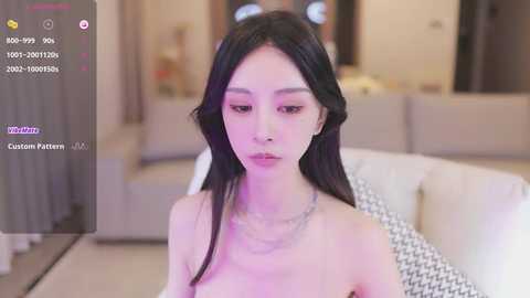 Media: Video of an East Asian woman with long black hair, light skin, and a slender physique, wearing a necklace, sitting on a white couch in a modern living room.